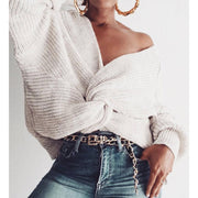 Close-up view a model wearing the Aries Sweater in milk white featuring a distinctive twisted front knot design. The chunky knit sweater is paired with gold bamboo earrings, a chain belt, and jeans. Worn off-shoulder with a wrap-style closure that creates a cropped silhouette. Product photography against a white background showcasing the sweater's chunky rib knit texture, soft yarns, relaxed fit, and versatile styling.