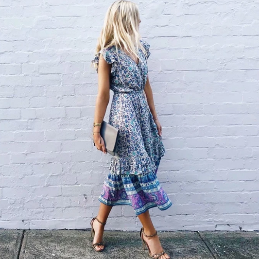 Nala Midi Dress from Shop The Kei - Blue and purple floral boho midi dress with flounce hem, worn by a blonde woman standing against a white brick wall, paired with strappy nude heels and a clutch