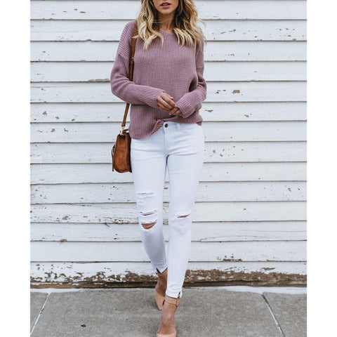 Full length front view of a model wearing the dusty purple Aries Sweater from Shop The Kei worn with its twist in an open back style. The chunky knit sweater is styled with white jeans, strappy suede heels, and a tan purse. Showcasing the sweater's casual and refreshed styling.