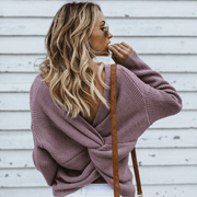 Back view of a model wearing the dusty purple colored Aries Sweater from Shop The Kei worn with its twist in an open back style. The chunky knit sweater is styled with white jeans, sunglasses, and a tan purse. Showcasing the sweater's relaxed and versatile styling.