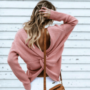 Back view of a model wearing the pink colored Aries Sweater from Shop The Kei worn with its twist in an open back style. The chunky knit sweater is styled with white jeans and a tan purse. Showcasing the sweater's relaxed and versatile styling.