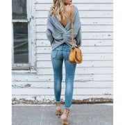Full length back view of a model wearing gray colored Aries Sweater from Shop The Kei worn with its twist in an open back style. The chunky knit sweater is styled with light blue jeans, a tan purse, and strappy heels. Showcasing the sweater's relaxed and versatile styling.