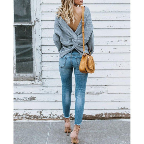 Full length back view of a model wearing gray colored Aries Sweater from Shop The Kei worn with its twist in an open back style. The chunky knit sweater is styled with light blue jeans, a tan purse, and strappy heels. Showcasing the sweater's relaxed and versatile styling.