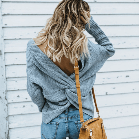Back view of a model wearing the gray colored Aries Sweater from Shop The Kei worn with its twist in an open back style. The chunky knit sweater is styled with light blue jeans and a tan purse. Showcasing the sweater's relaxed and versatile styling.