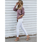 Full length side view of a model wearing the dusty purple Aries Sweater from Shop The Kei worn with its twist in an open back style. The chunky knit sweater is styled with white jeans, strappy suede heels, and a tan purse. Showcasing the sweater's refreshed keen style