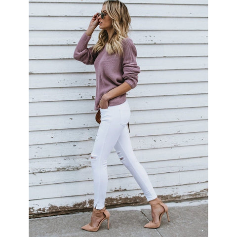 Full length side view of a model wearing the dusty purple Aries Sweater from Shop The Kei worn with its twist in an open back style. The chunky knit sweater is styled with white jeans, strappy suede heels, and a tan purse. Showcasing the sweater's refreshed keen style