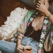 Model sits wearing Nyma Kimono from Shop The Kei swim coverup collection with a lacy black tank and jeans. Bohemian-style Nyma Kimono with open front, waist sash, wing sleeves, and ankle-length floral print rayon fabric. Elegant and flowy