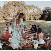 Model stands at a picnic with friends wearing Nyma Kimono from Shop The Kei swim coverup collection with a large stone ring. Bohemian-style Nyma Kimono with open front, waist sash, wing sleeves, and ankle-length floral print rayon fabric. Elegant and flowy cover up.