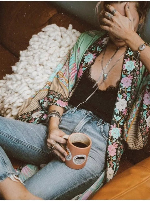 Model sits wearing Nyma Kimono from Shop The Kei swim coverup collection with a lacy black tank and jeans. Bohemian-style Nyma Kimono with open front, waist sash, wing sleeves, and ankle-length floral print rayon fabric. Elegant and flowy