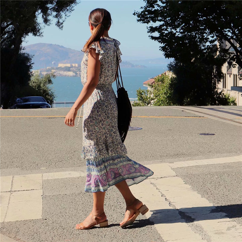 Nala Midi Dress from Shop The Kei - Back view of blue and purple floral boho midi dress with flounce hem and ruffle sleeves, worn by a brunette woman crossing the street, paired with nude sandals and a fringe purse