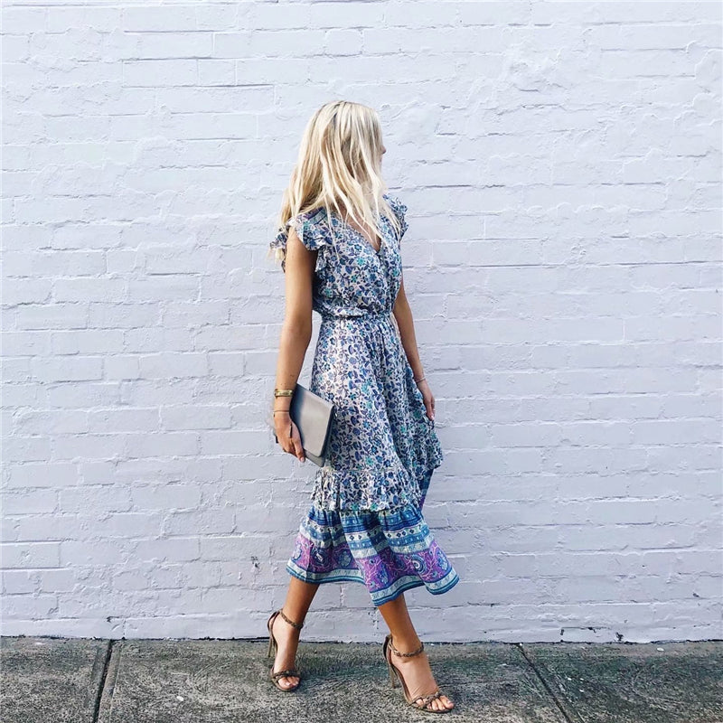 Nala Midi Dress from Shop The Kei - Blue and purple floral boho midi dress with flounce hem, worn by a blonde woman standing against a white brick wall, paired with strappy nude heels and a clutch