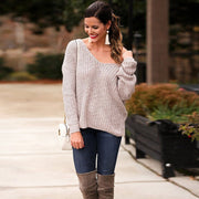Front view of a model in a garden landscape wearing oat colored Aries Sweater from Shop The Kei worn with its twist in an open back style. Drop shoulders worn loosely with a wide v neckline. The chunky knit sweater is styled with blue jeans, white purse, and tan boots. Showcasing the sweater's relaxed and versatile styling.