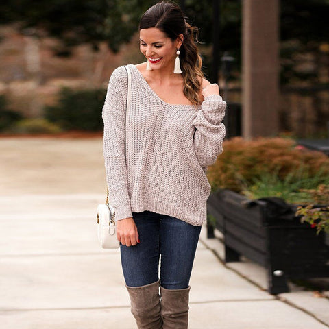 Front view of a model in a garden landscape wearing oat colored Aries Sweater from Shop The Kei worn with its twist in an open back style. Drop shoulders worn loosely with a wide v neckline. The chunky knit sweater is styled with blue jeans, white purse, and tan boots. Showcasing the sweater's relaxed and versatile styling.