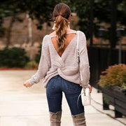 Back view of a model in a garden landscape wearing oat colored Aries Sweater from Shop The Kei worn with its twist in an open back style. The chunky knit sweater is styled with blue jeans, white purse, and tan boots. Showcasing the sweater's relaxed and versatile styling.