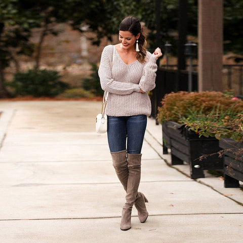 Full length front view of a model in a garden landscape wearing oat colored Aries Sweater from Shop The Kei worn with its twist in an open back style. Drop shoulders worn loosely with a wide v neckline. The chunky knit sweater is styled with blue jeans, white purse, and tan boots. Showcasing the sweater's relaxed and versatile styling.