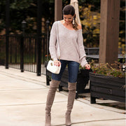 Full length front view of a model wearing oat colored Aries Sweater from Shop The Kei worn with its long full sleeves and drop shoulders worn loosely with a wide v neckline. The chunky knit sweater is styled with blue jeans, white purse, and tan boots. Showcasing the sweater's relaxed and versatile styling.